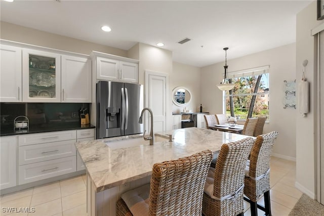 kitchen featuring stainless steel fridge, white cabinetry, hanging light fixtures, light stone countertops, and an island with sink