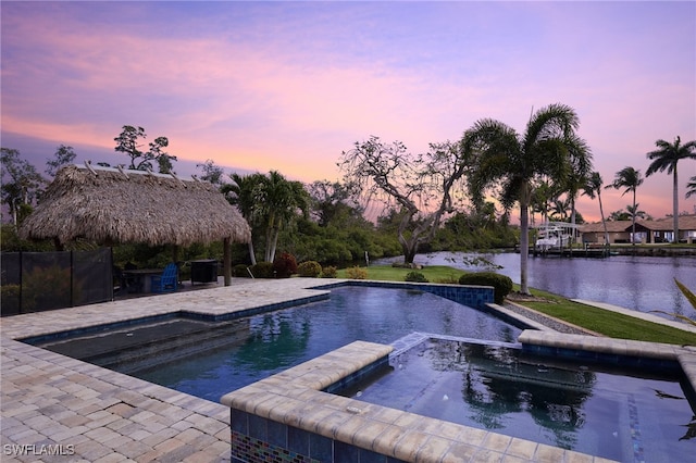 pool at dusk with a pool with connected hot tub, a patio area, and a water view