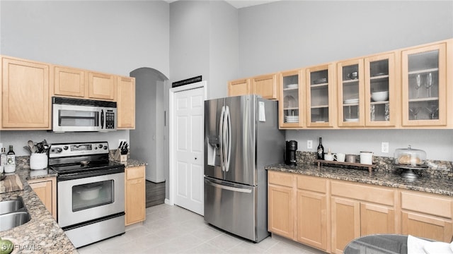 kitchen featuring a towering ceiling, appliances with stainless steel finishes, light brown cabinets, and dark stone counters