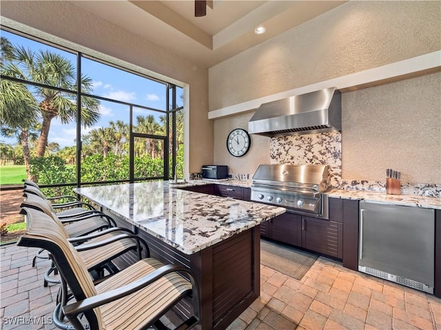 kitchen with wall chimney exhaust hood, a kitchen bar, refrigerator, and light stone countertops