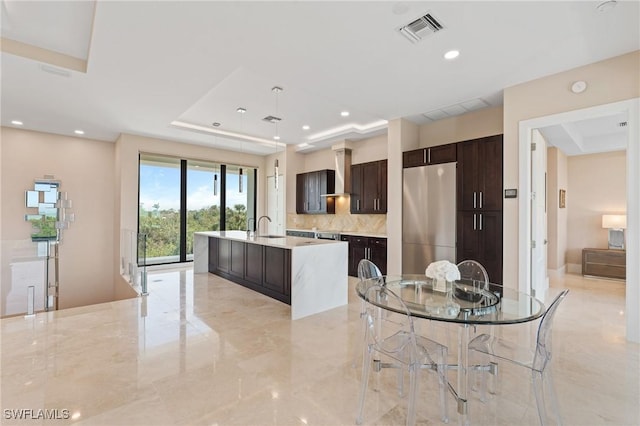 kitchen with stainless steel fridge, dark brown cabinetry, an island with sink, a raised ceiling, and wall chimney exhaust hood