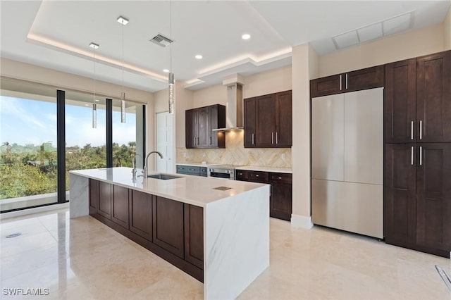 kitchen with wall chimney exhaust hood, sink, hanging light fixtures, a raised ceiling, and an island with sink