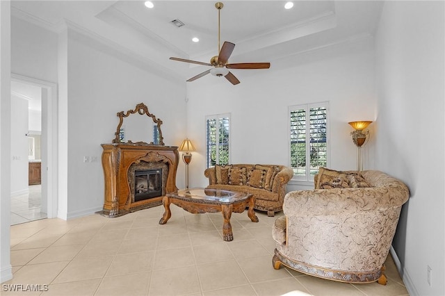 living area featuring a raised ceiling, a glass covered fireplace, ceiling fan, crown molding, and light tile patterned flooring