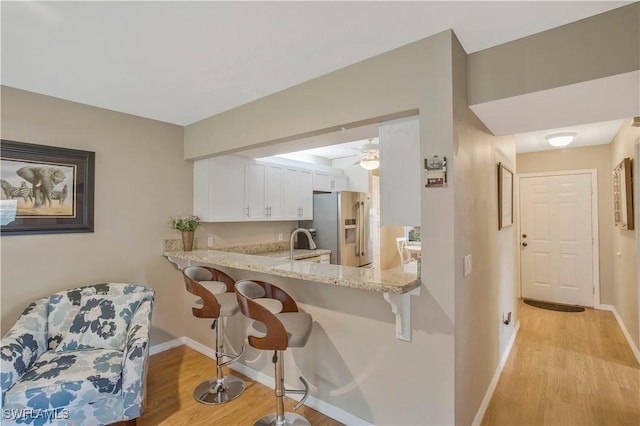 kitchen featuring light wood-style flooring, stainless steel refrigerator with ice dispenser, light stone counters, a kitchen breakfast bar, and white cabinetry