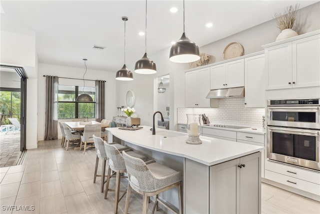 kitchen with white cabinetry, backsplash, an island with sink, decorative light fixtures, and stainless steel double oven