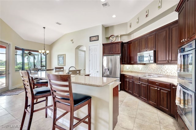 kitchen featuring a center island with sink, lofted ceiling, stainless steel appliances, decorative light fixtures, and light stone counters