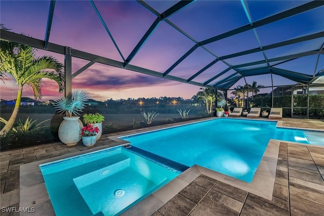 pool at dusk featuring glass enclosure, a patio area, and a pool with connected hot tub