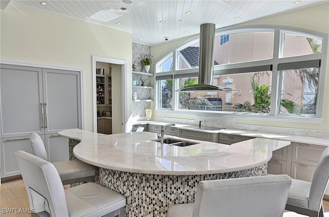 kitchen with a center island with sink, gray cabinets, island exhaust hood, light stone counters, and a sink