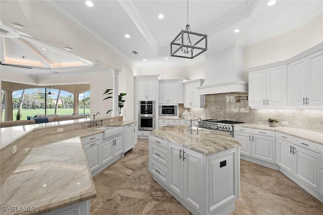 kitchen with a tray ceiling, custom exhaust hood, stainless steel appliances, backsplash, and a kitchen island with sink