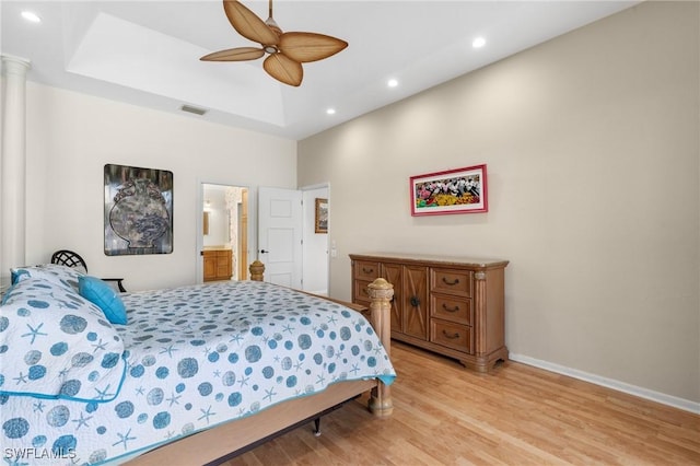 bedroom with ornate columns, ceiling fan, a raised ceiling, ensuite bath, and light hardwood / wood-style flooring