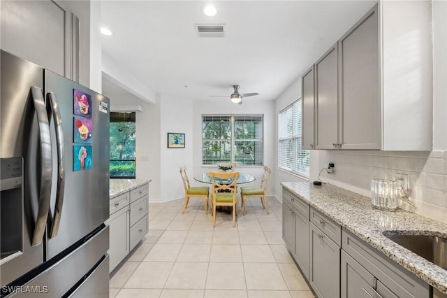 kitchen with light tile patterned floors, gray cabinets, light stone countertops, stainless steel fridge with ice dispenser, and decorative backsplash