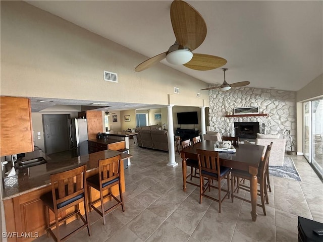 dining space featuring visible vents, a ceiling fan, a stone fireplace, ornate columns, and high vaulted ceiling