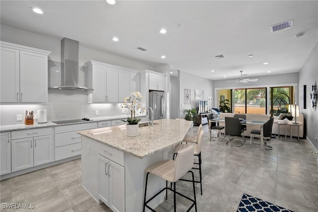 kitchen featuring black electric stovetop, a kitchen island with sink, visible vents, freestanding refrigerator, and wall chimney exhaust hood