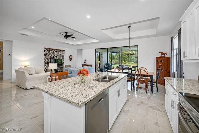 kitchen featuring an island with sink, sink, hanging light fixtures, stainless steel dishwasher, and a tray ceiling