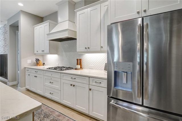 kitchen featuring stainless steel appliances, custom range hood, backsplash, light wood-style floors, and white cabinetry