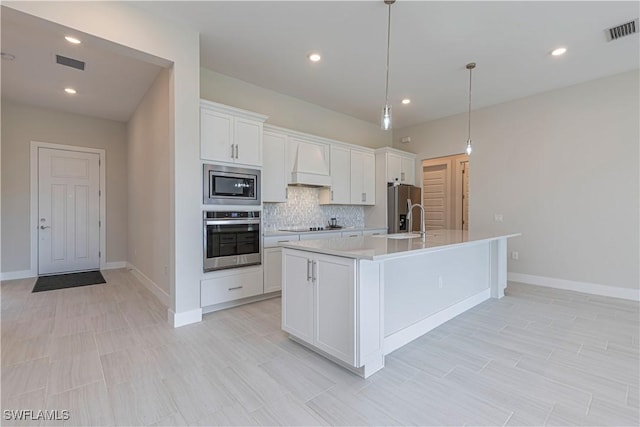 kitchen featuring an island with sink, stainless steel appliances, premium range hood, white cabinets, and pendant lighting