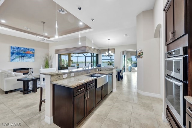 kitchen featuring appliances with stainless steel finishes, hanging light fixtures, a tray ceiling, light stone countertops, and a center island with sink