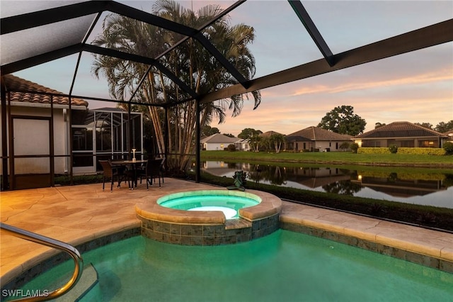 pool at dusk with an in ground hot tub, a water view, glass enclosure, and a patio area