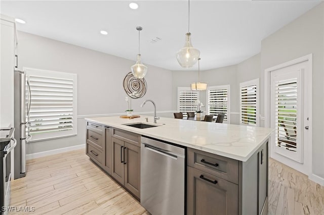 kitchen with sink, light stone counters, decorative light fixtures, appliances with stainless steel finishes, and an island with sink