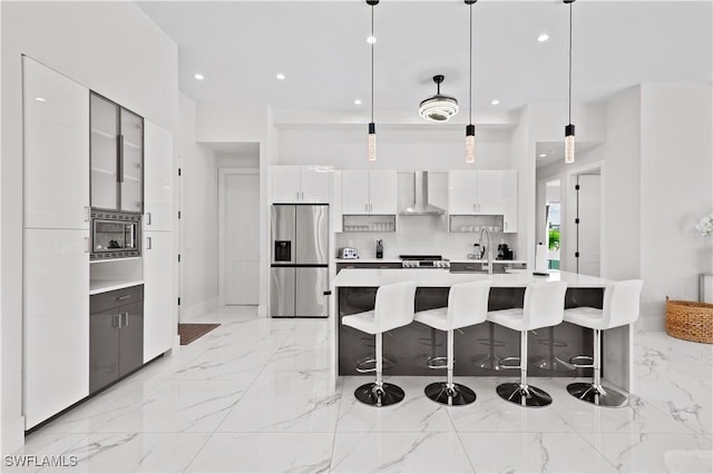 kitchen featuring pendant lighting, an island with sink, white cabinets, stainless steel appliances, and wall chimney range hood