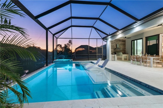 pool at dusk featuring a patio, an outdoor kitchen, exterior bar, ceiling fan, and glass enclosure