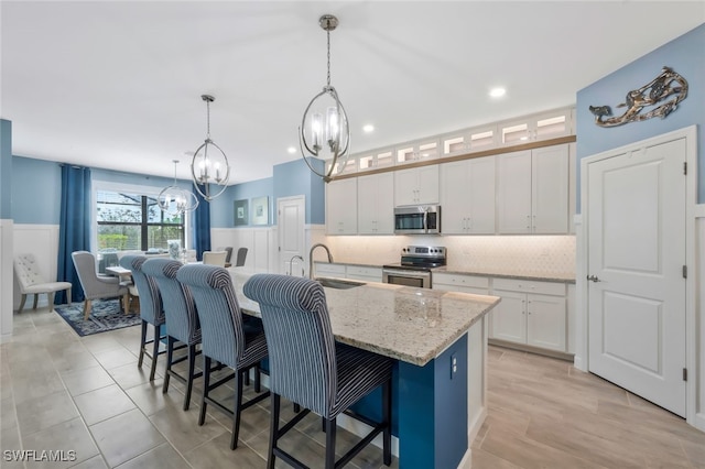 kitchen with white cabinets, an island with sink, hanging light fixtures, light stone countertops, and stainless steel appliances