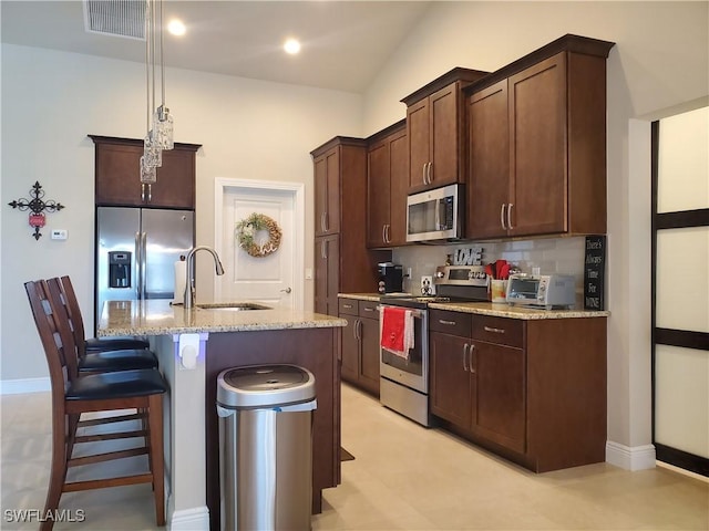 kitchen featuring sink, appliances with stainless steel finishes, hanging light fixtures, light stone counters, and an island with sink