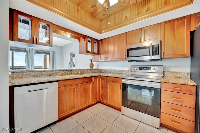 kitchen with light stone counters, a tray ceiling, stainless steel appliances, glass insert cabinets, and a sink