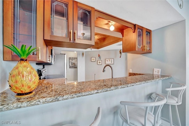 kitchen featuring brown cabinets, stove, glass insert cabinets, light stone countertops, and a peninsula