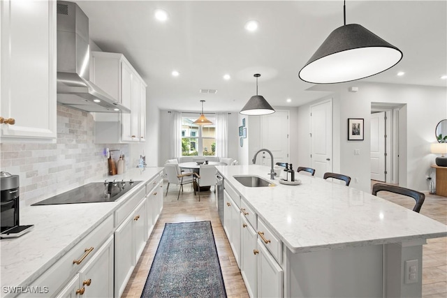 kitchen featuring white cabinets, an island with sink, black electric cooktop, wall chimney range hood, and a sink