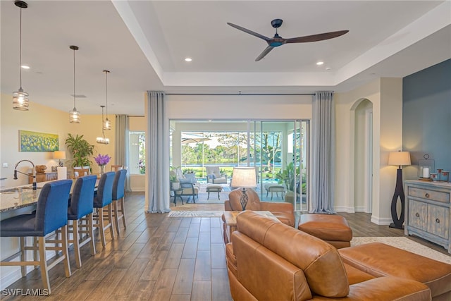 living area with a ceiling fan, a tray ceiling, and dark wood-type flooring