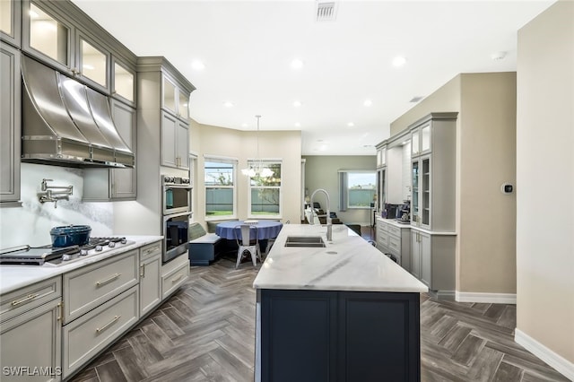 kitchen with glass insert cabinets, appliances with stainless steel finishes, a kitchen island with sink, a sink, and exhaust hood