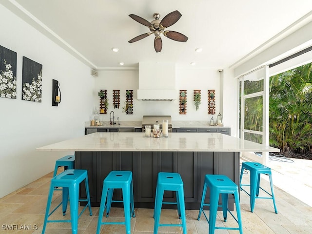 kitchen with a ceiling fan, a spacious island, stone tile flooring, premium range hood, and a sink