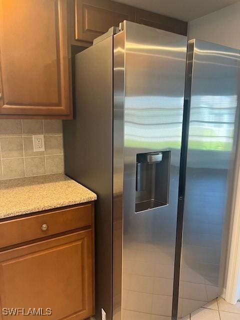 kitchen with light tile patterned flooring, stainless steel refrigerator with ice dispenser, light stone countertops, tasteful backsplash, and brown cabinetry