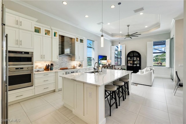 kitchen featuring a sink, a center island with sink, wall chimney exhaust hood, glass insert cabinets, and pendant lighting