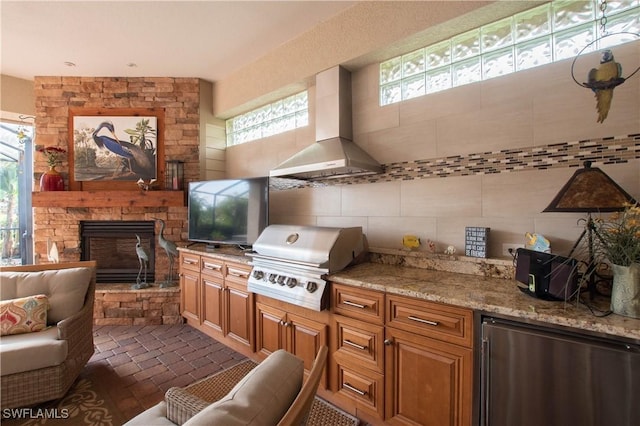 kitchen featuring a stone fireplace, light stone counters, wall chimney range hood, backsplash, and stainless steel fridge