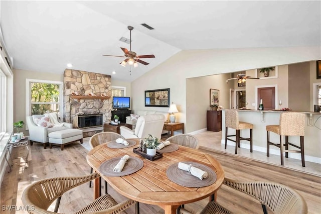 dining area featuring light wood finished floors, visible vents, ceiling fan, vaulted ceiling, and a stone fireplace