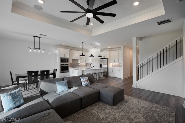 living room featuring a tray ceiling, crown molding, a ceiling fan, visible vents, and dark wood-type flooring