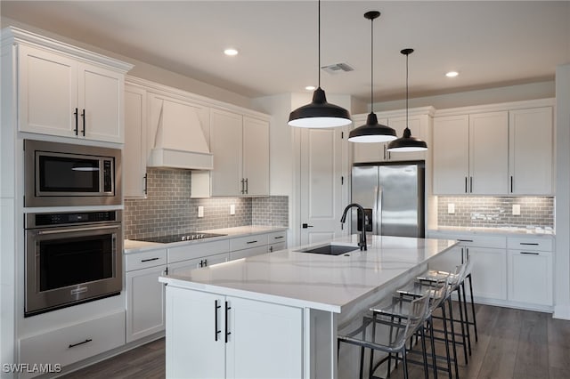 kitchen with a center island with sink, premium range hood, stainless steel appliances, sink, and white cabinetry