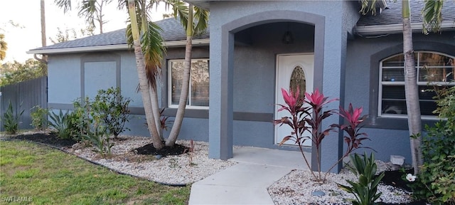 doorway to property with stucco siding, fence, and a shingled roof