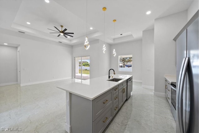 kitchen featuring pendant lighting, sink, appliances with stainless steel finishes, a tray ceiling, and a center island with sink