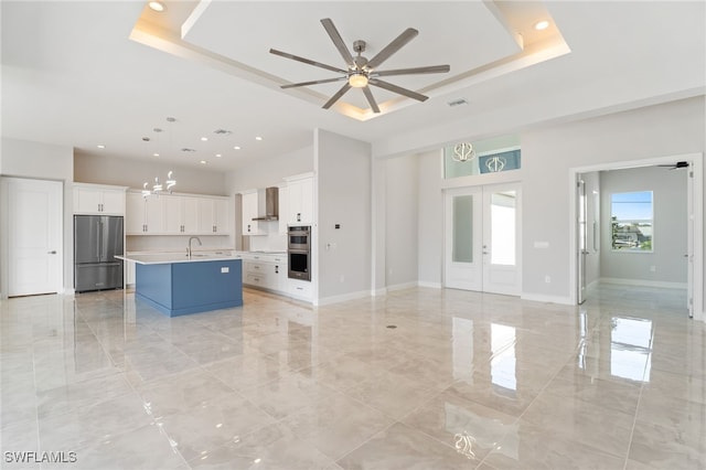 kitchen with appliances with stainless steel finishes, a center island with sink, wall chimney range hood, a raised ceiling, and white cabinetry