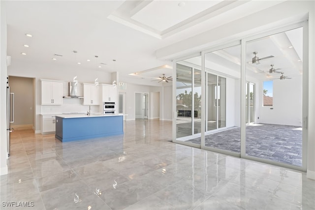 kitchen with hanging light fixtures, ceiling fan, wall chimney exhaust hood, white cabinets, and an island with sink