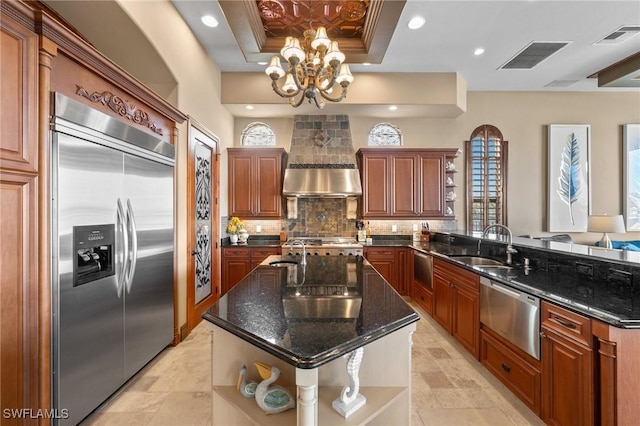 kitchen with a tray ceiling, dark stone counters, stainless steel built in refrigerator, ventilation hood, and an island with sink