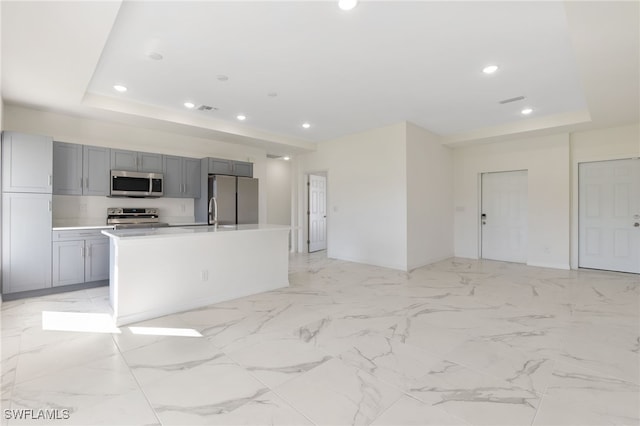 kitchen featuring a kitchen island with sink, a tray ceiling, gray cabinets, and appliances with stainless steel finishes