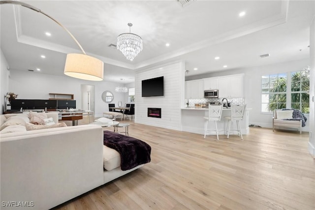 living room with light wood-type flooring, ornamental molding, an inviting chandelier, and a raised ceiling