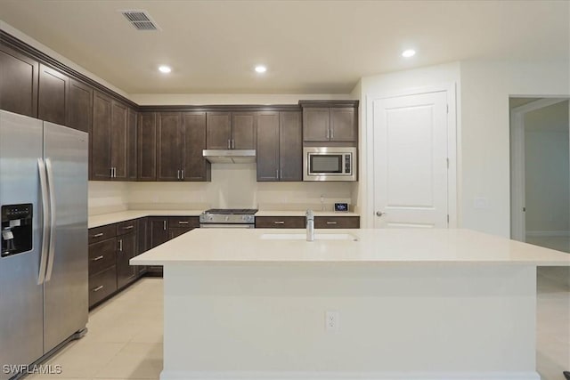 kitchen with appliances with stainless steel finishes, dark brown cabinets, an island with sink, light tile patterned flooring, and sink