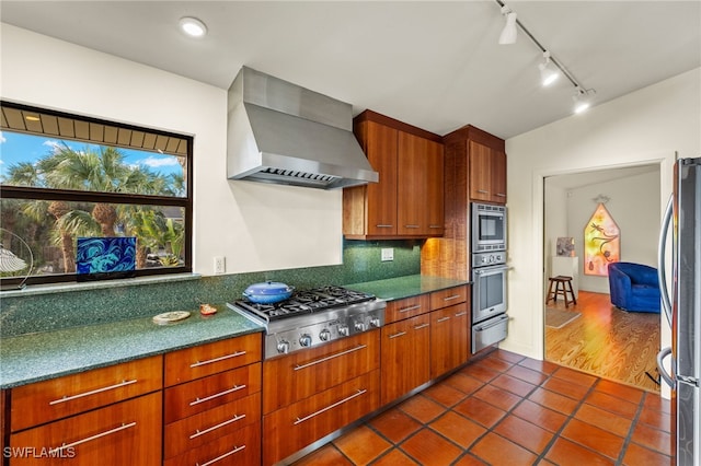 kitchen with stainless steel appliances, backsplash, dark tile patterned flooring, track lighting, and wall chimney range hood