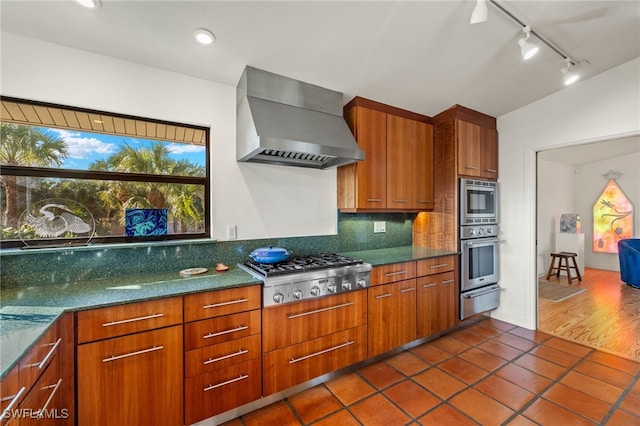 kitchen with appliances with stainless steel finishes, tasteful backsplash, wall chimney range hood, dark tile patterned floors, and dark stone countertops