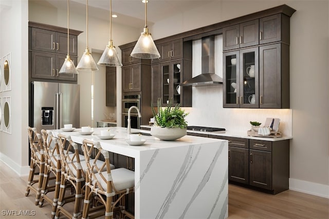 kitchen with wall chimney range hood, dark brown cabinets, a center island with sink, and appliances with stainless steel finishes